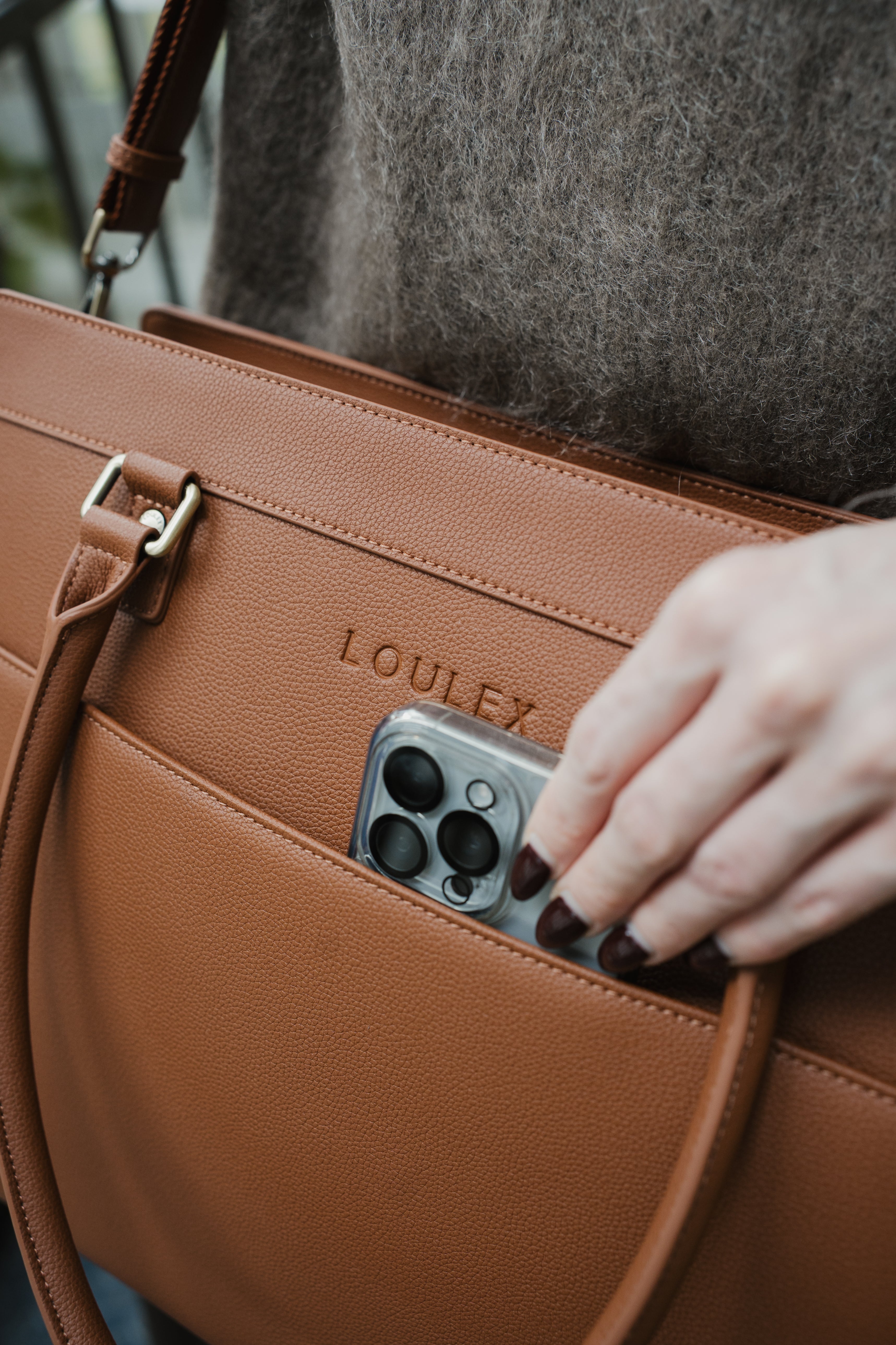 Brown leather bag with 'LOULEX' branding, held by a person with a phone in the pocket.