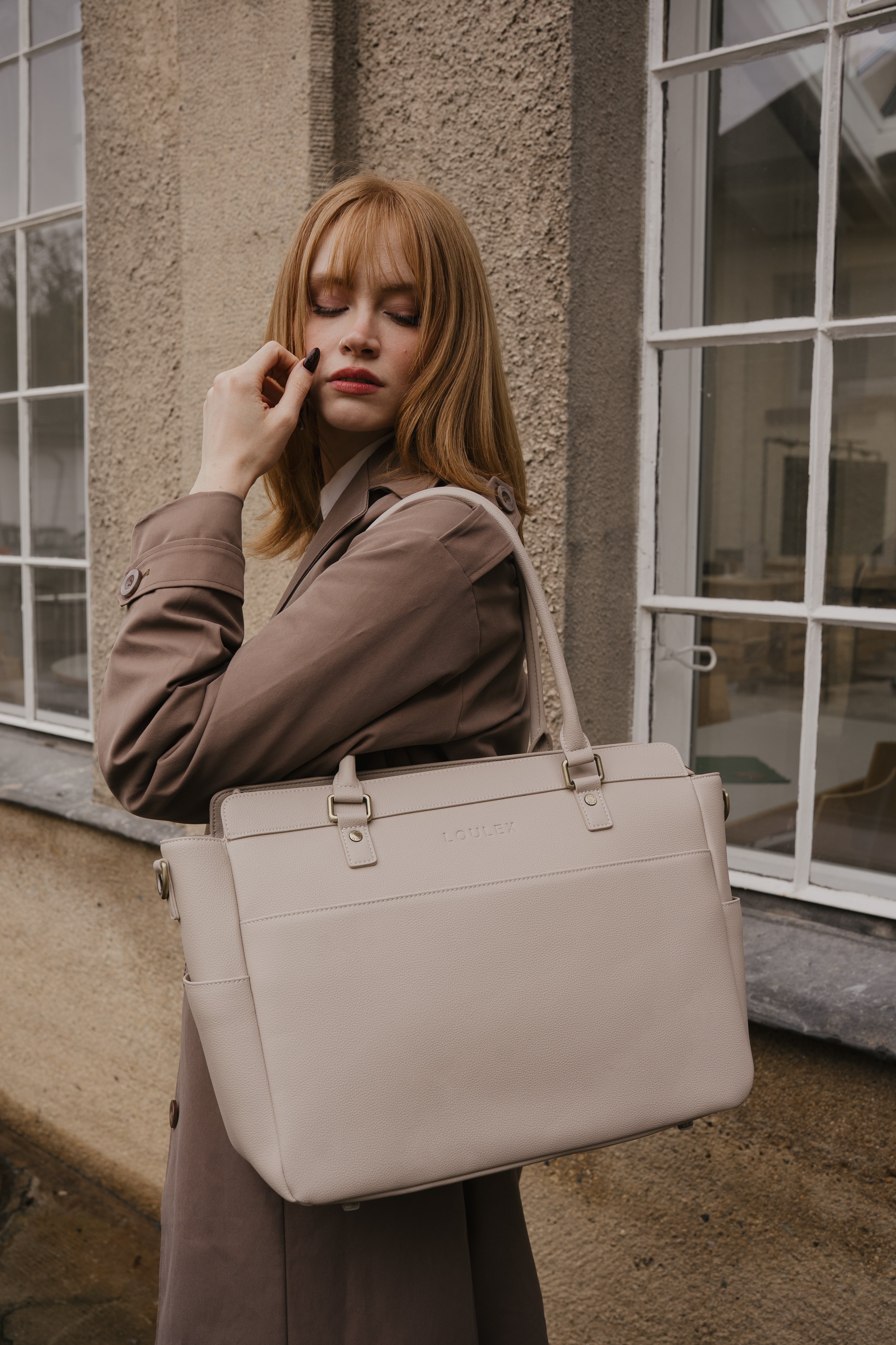 Woman holding a beige handbag in front of a building with large windows.