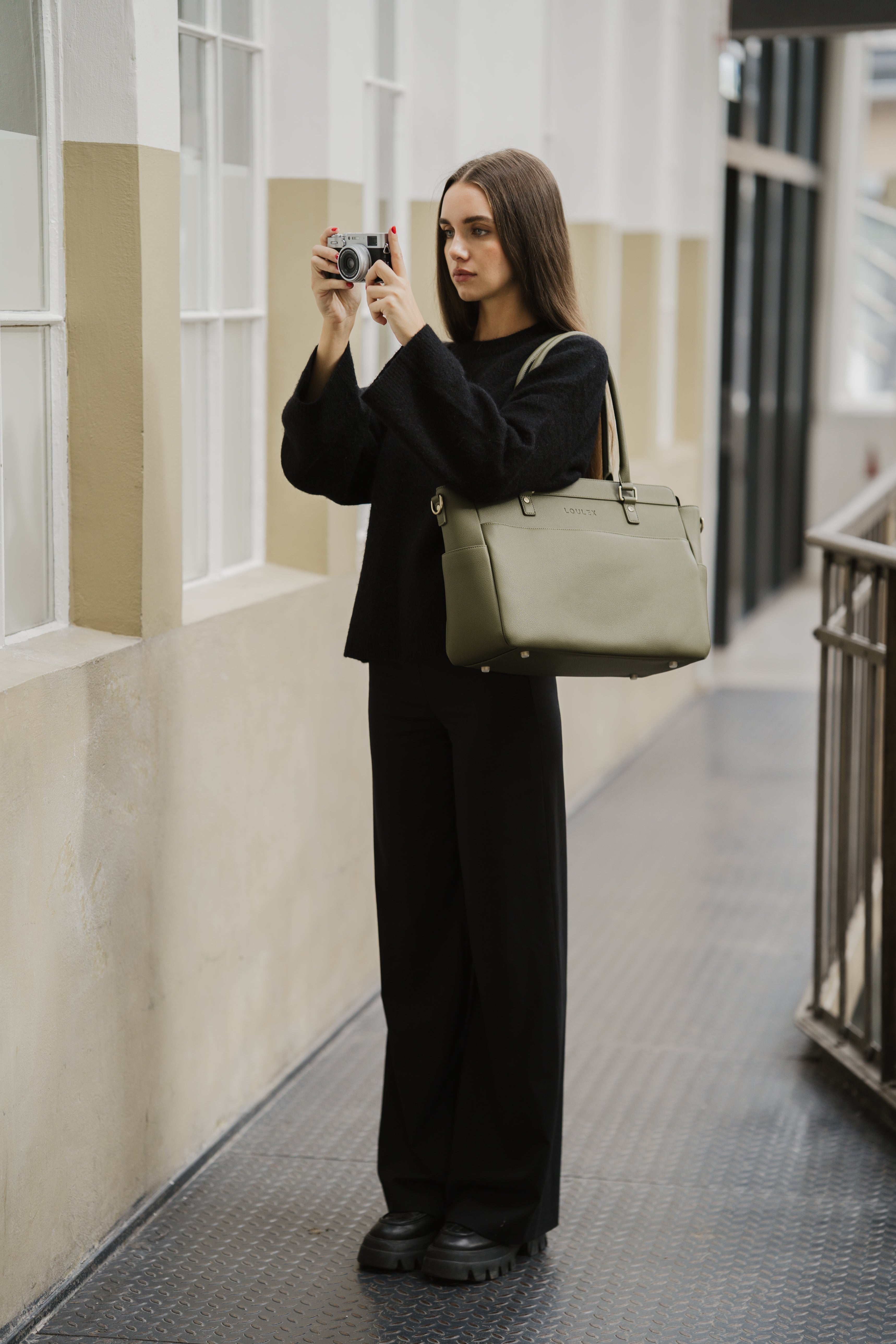 Person in a black outfit holding a camera and a green bag in an indoor setting.