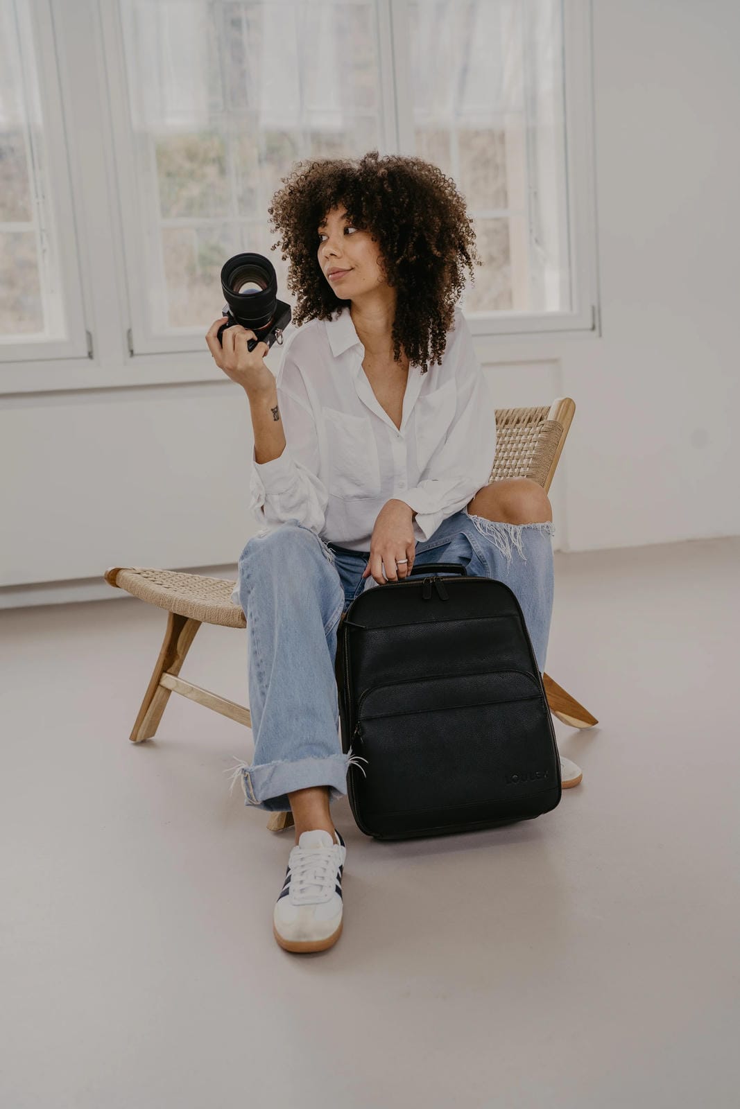 Woman holding a camera and a black backpack in a minimalistic room.