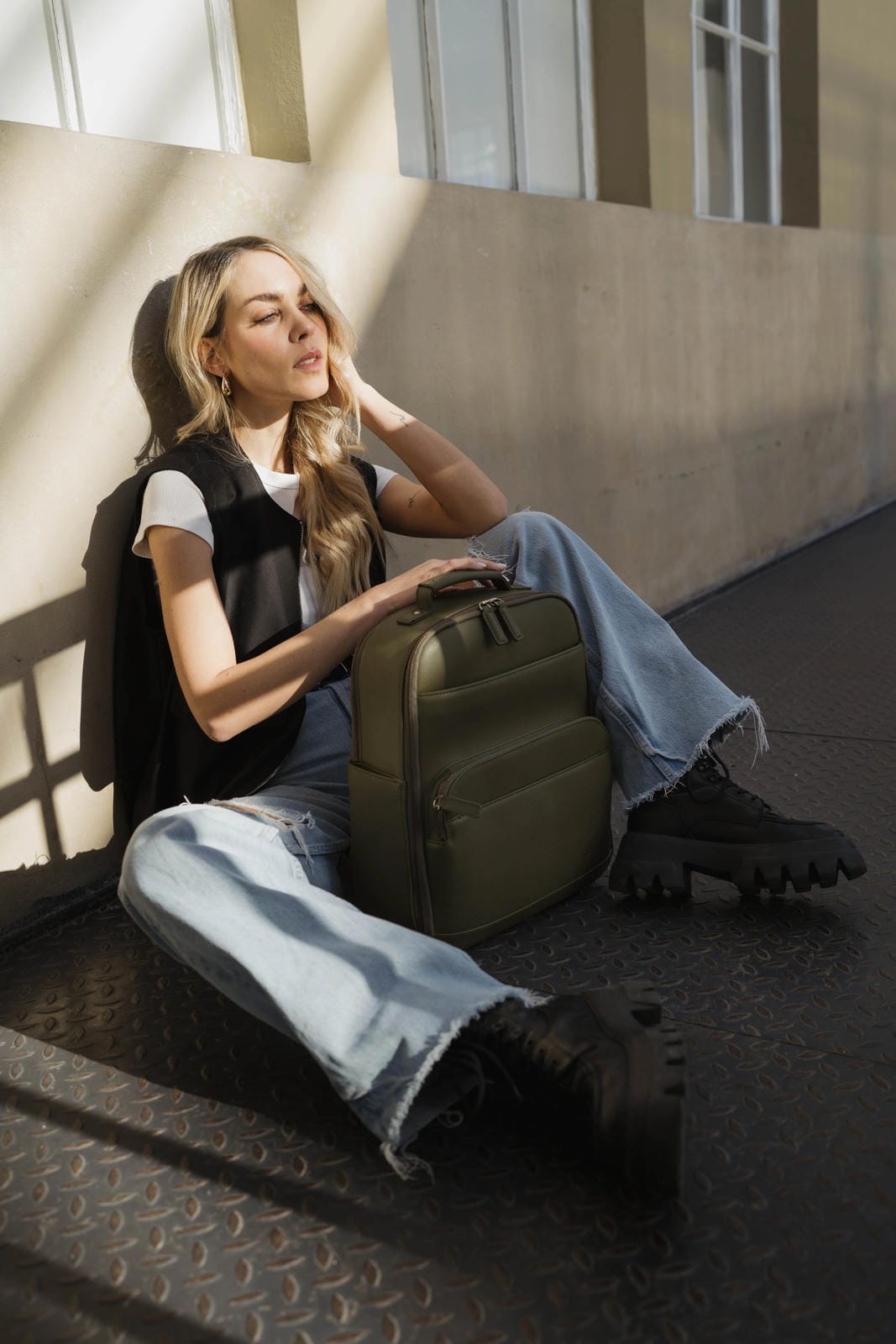 Woman sitting on a bench with a green backpack, wearing a black jacket and blue jeans.