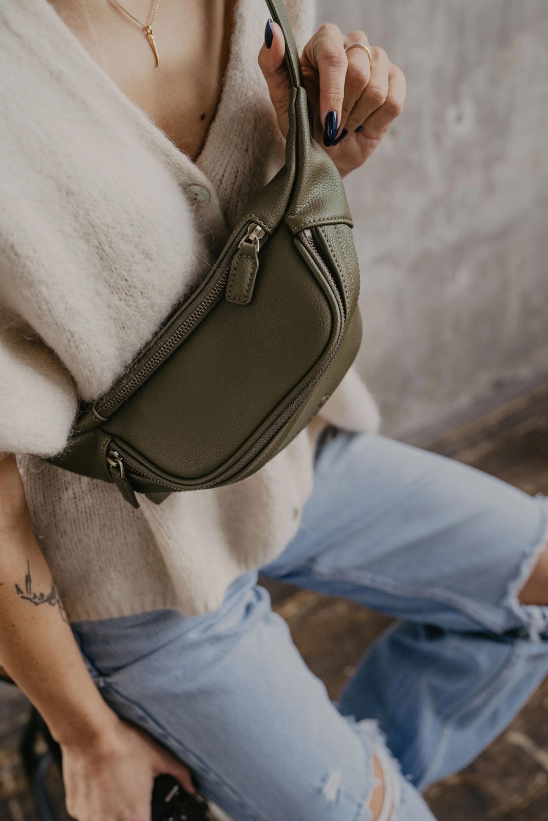 Person wearing a beige sweater and blue jeans with a green waist bag.