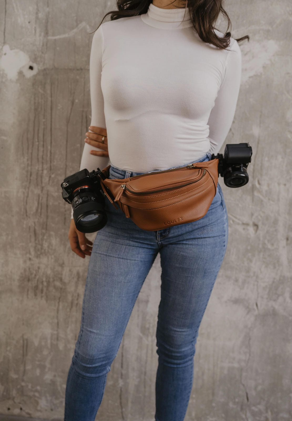 Person wearing a white turtleneck, blue jeans, and a brown fanny pack with camera equipment against a concrete wall.