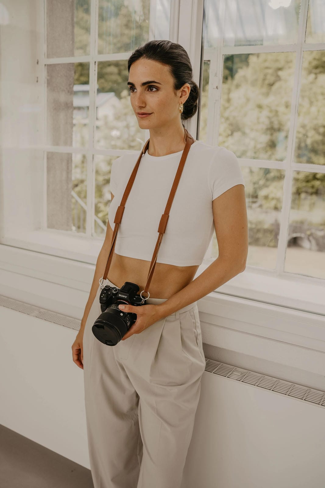 Woman holding a camera with a strap, standing in a room with large windows.
