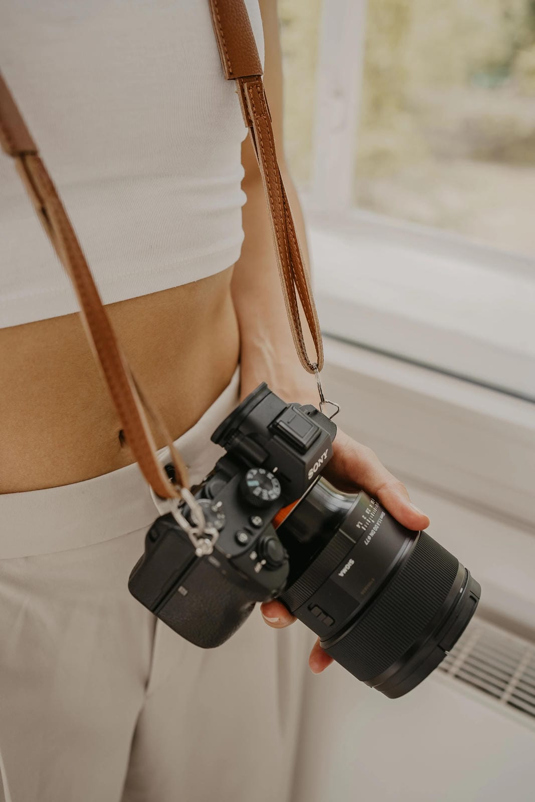 Person holding a camera with a strap, standing indoors near a window.