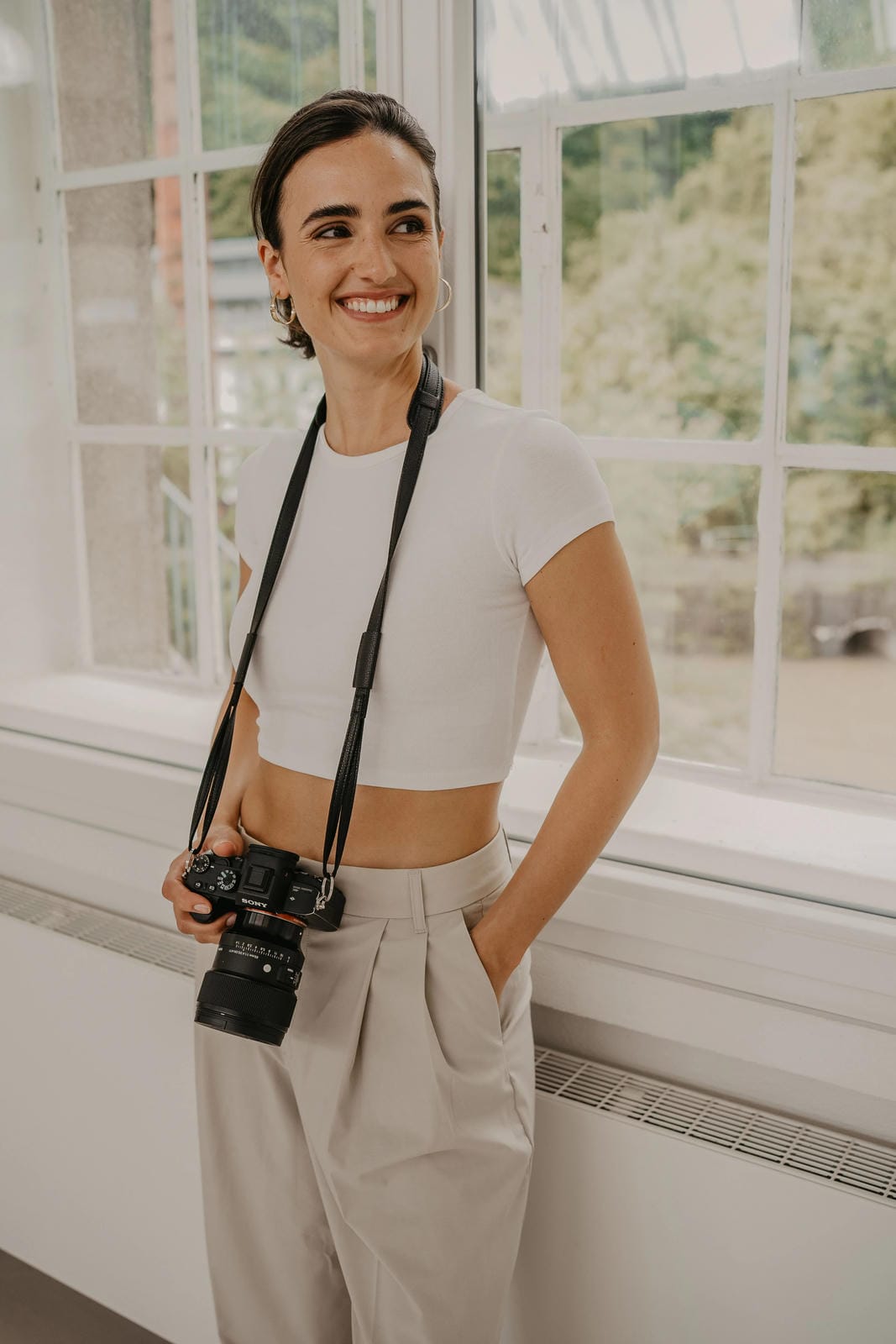 Woman holding a camera, standing in a room with large windows.