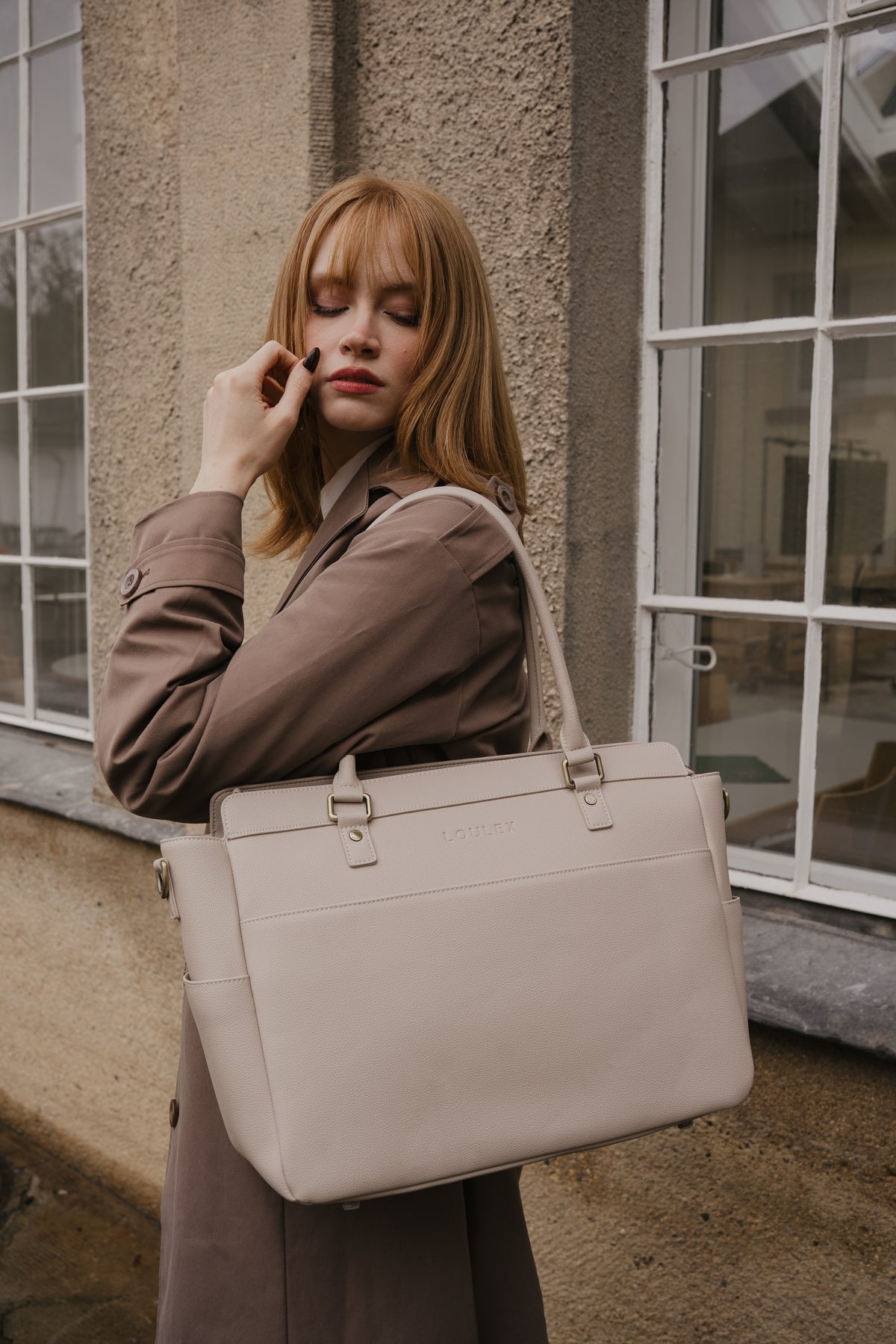 Woman holding a beige handbag in front of a building with large windows.