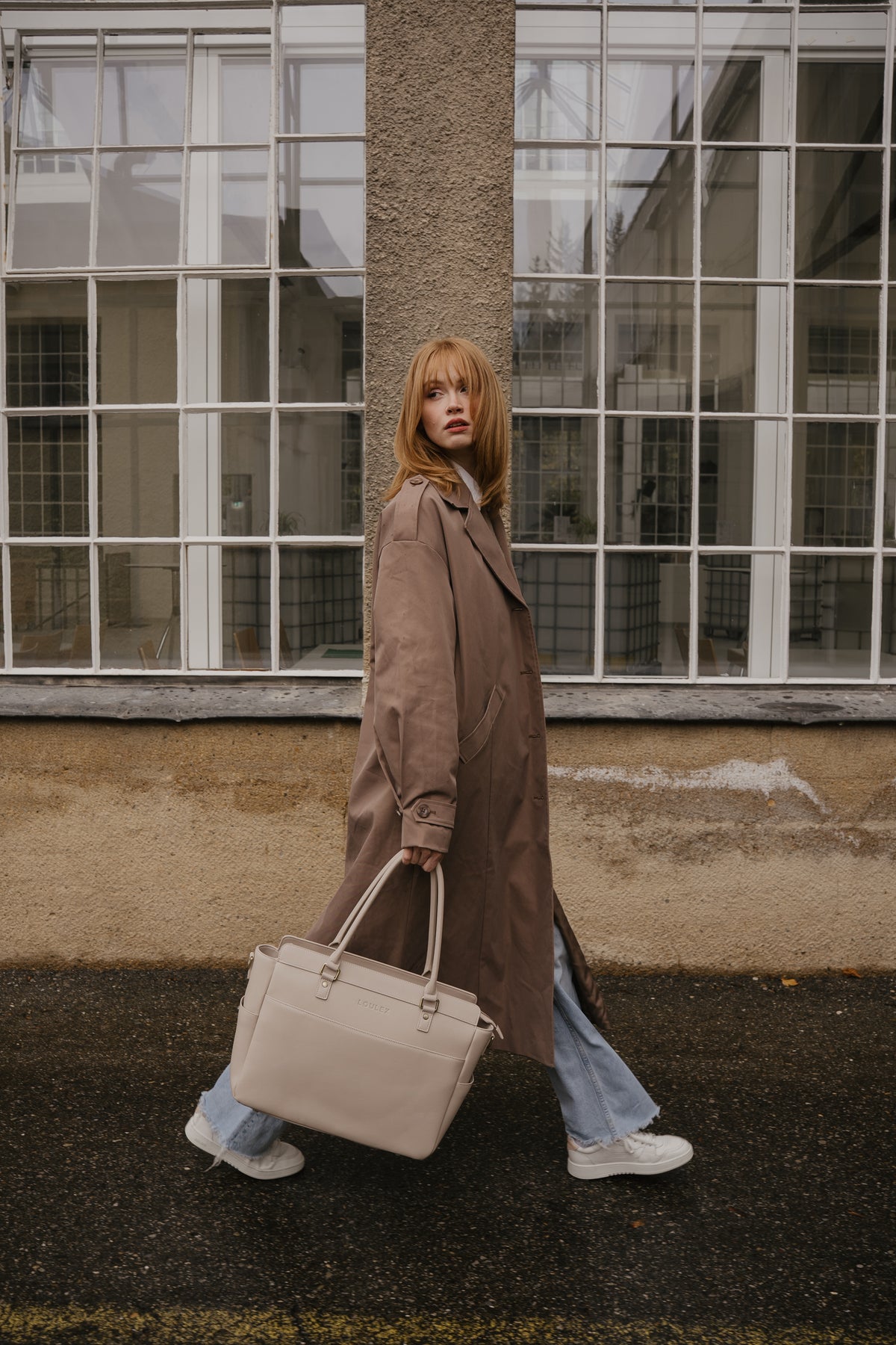 Woman in a long coat holding a beige bag, standing in front of a building with large windows.