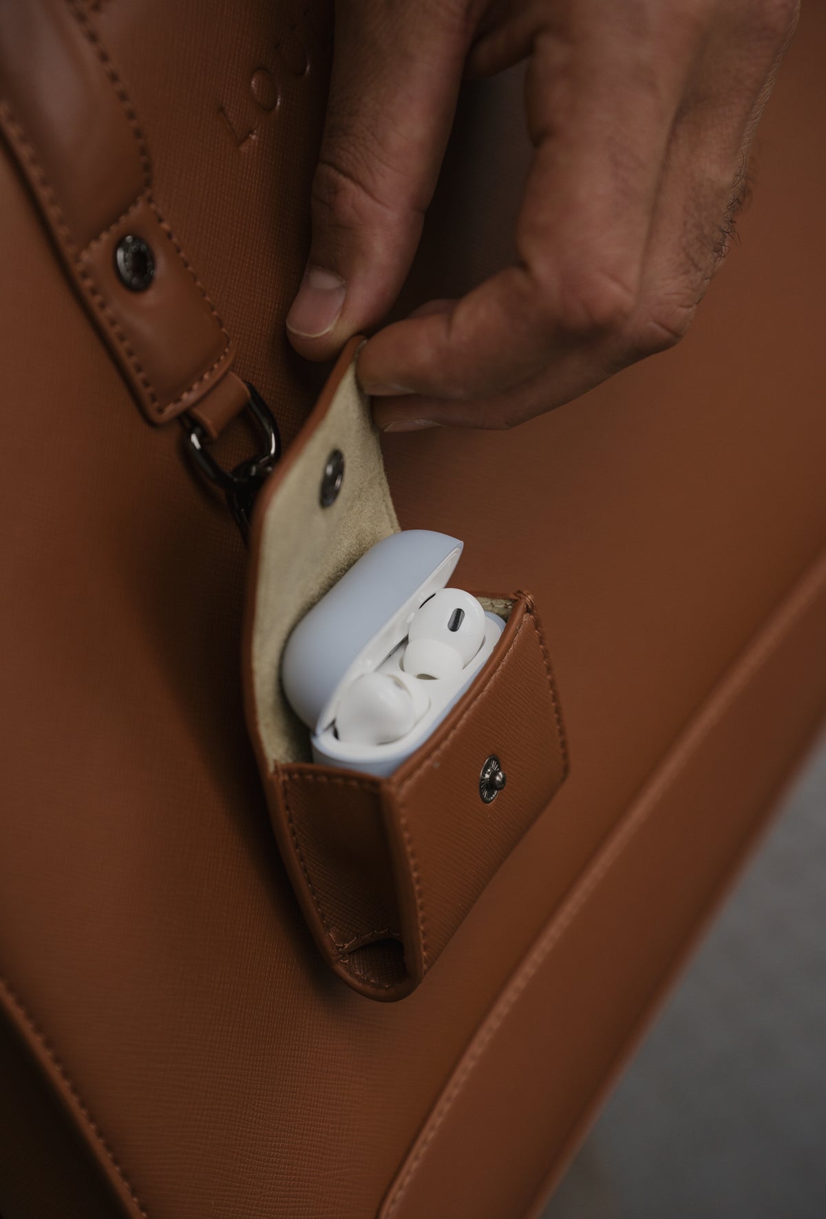 Brown leather case with a small pocket containing white earbuds, held by a hand.