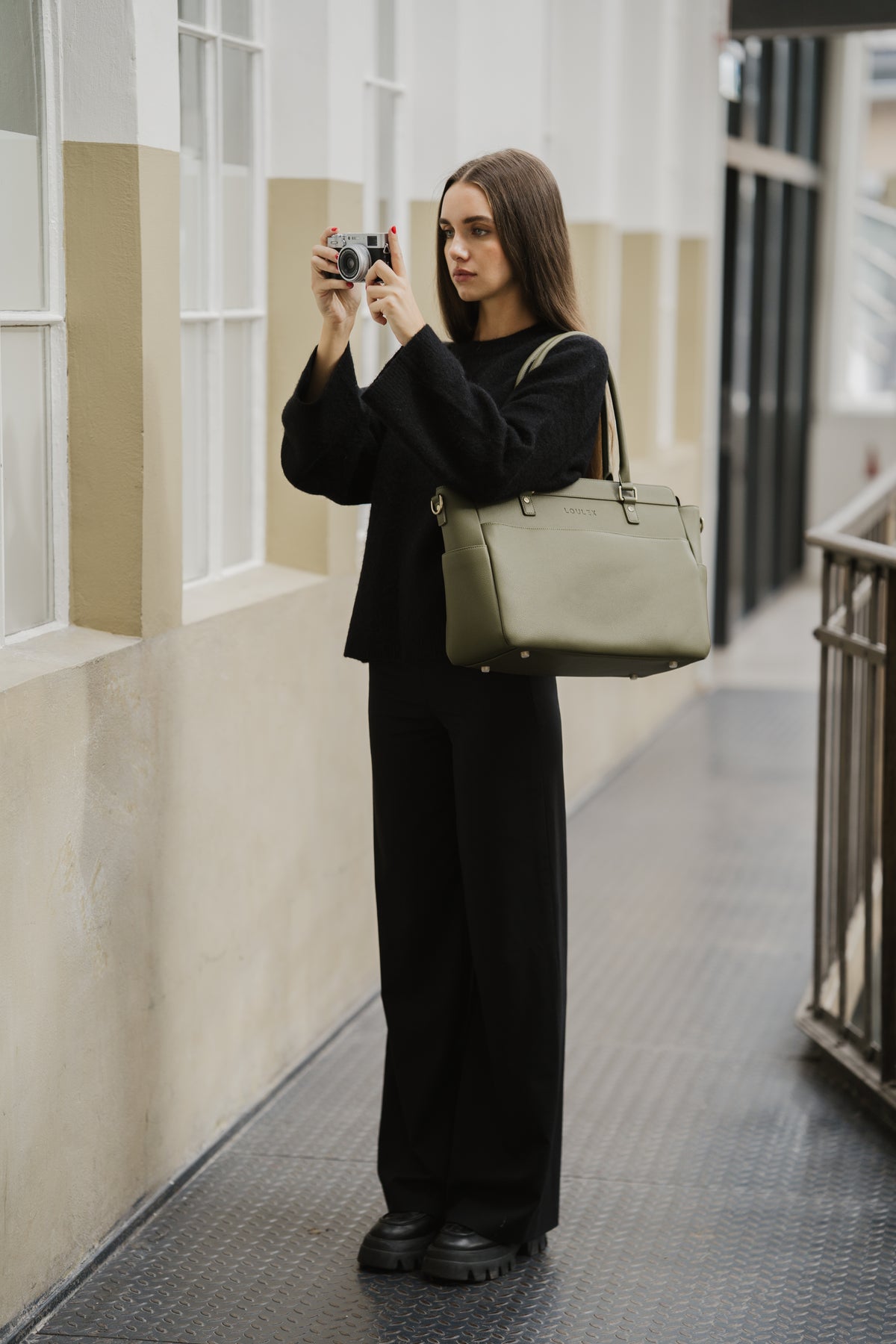 Person in a black outfit holding a camera and a green bag in an indoor setting.