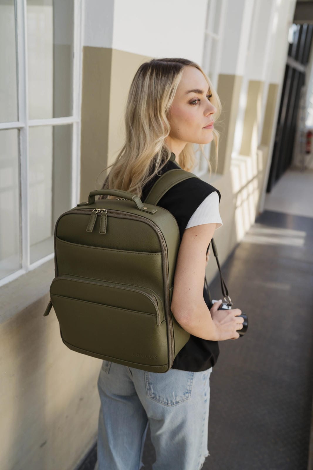 Woman wearing an olive green backpack and holding a camera, standing against a building wall.