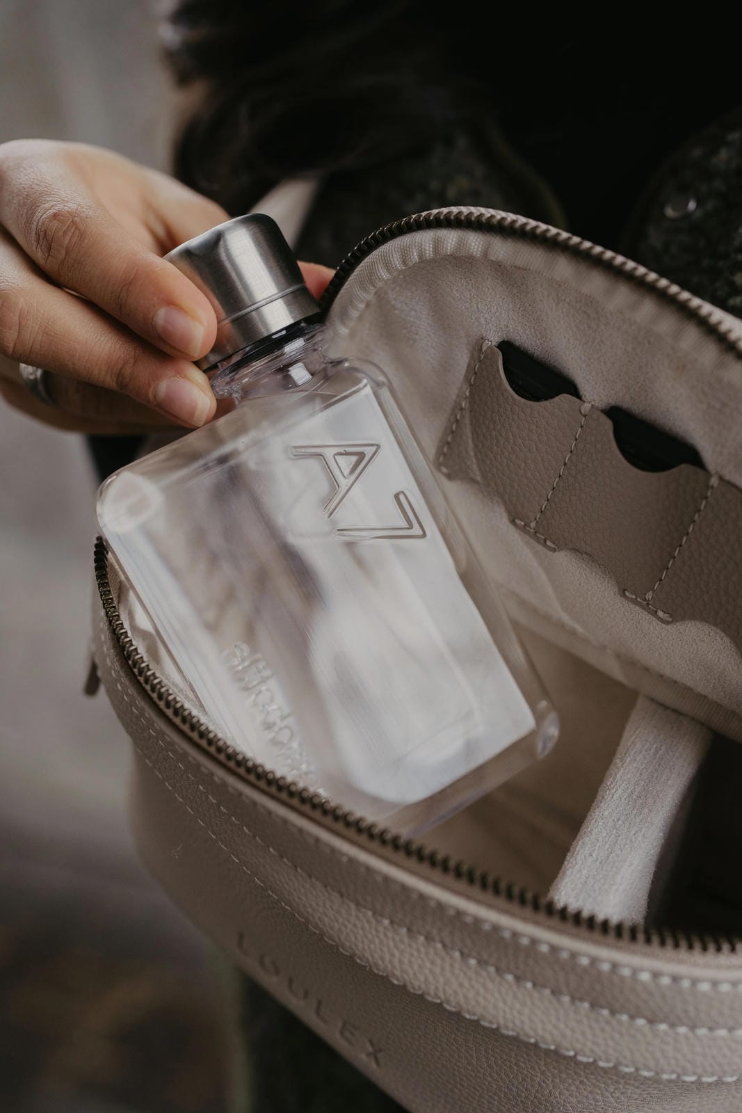 Person holding a clear glass bottle with a silver cap over an open beige bag.