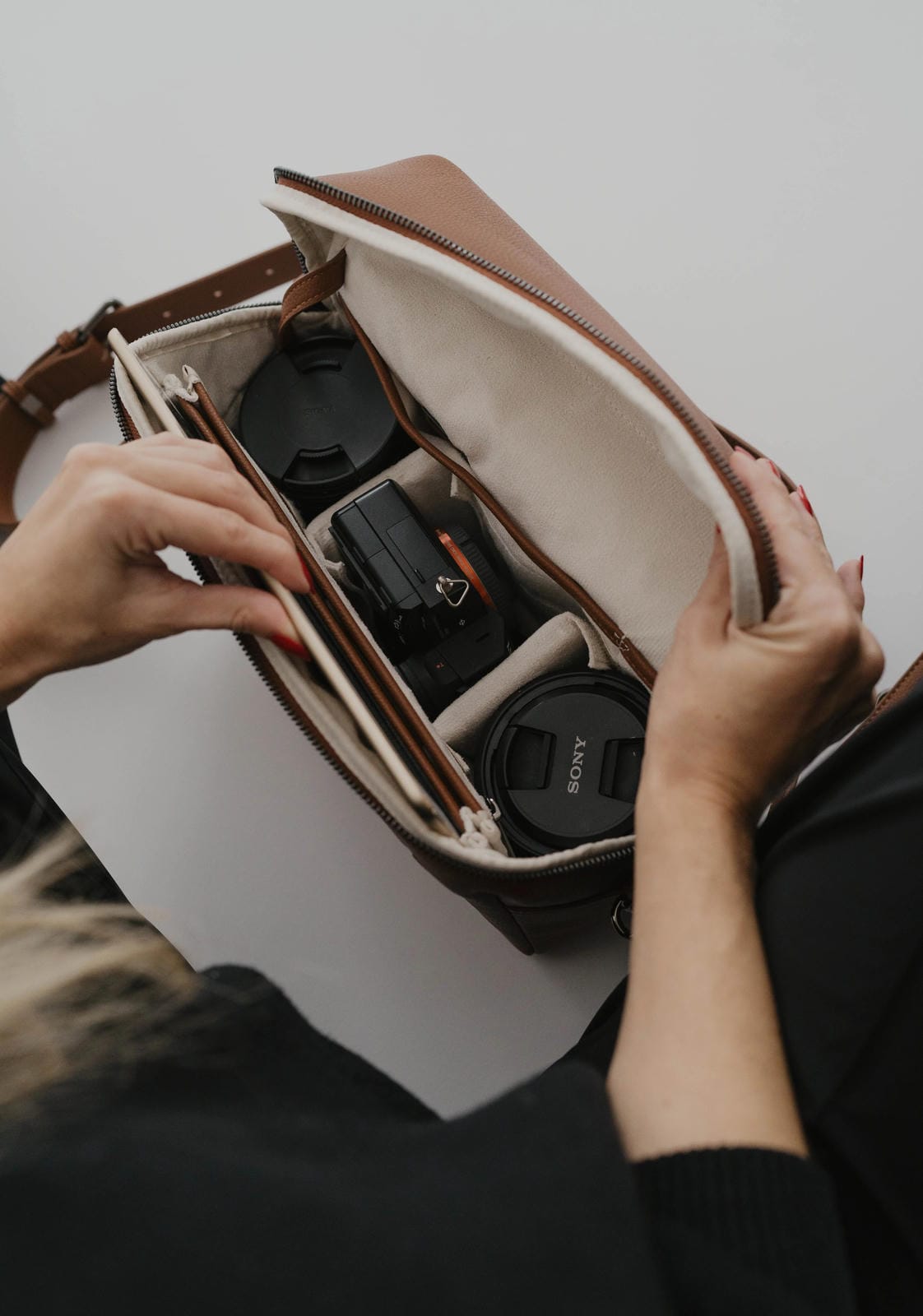 Person organizing camera equipment in a brown leather bag against a neutral background