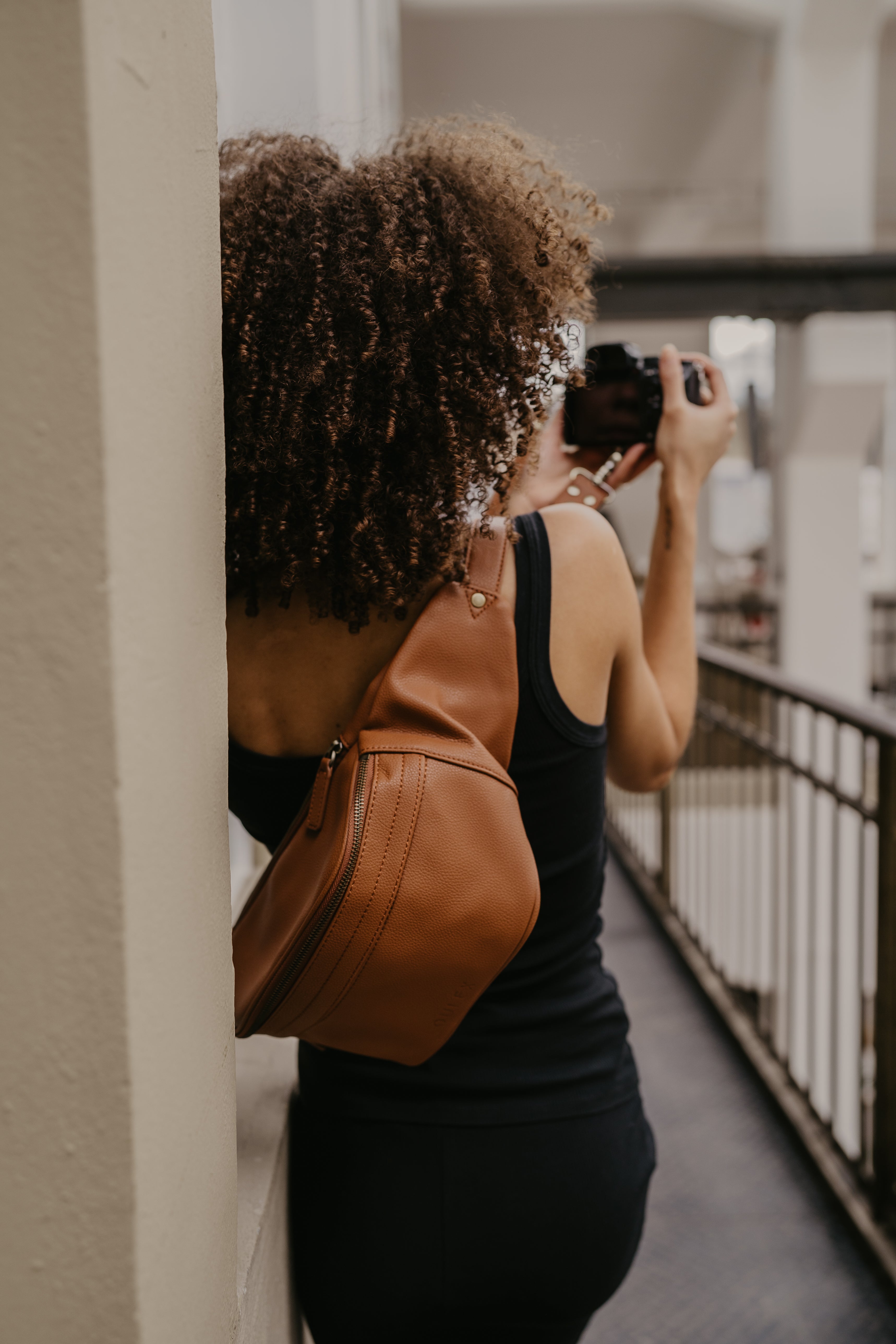 Woman in a grey knit dress wearing a loulex black leather photographer backpack while taking a photo in an industrial studio setting.