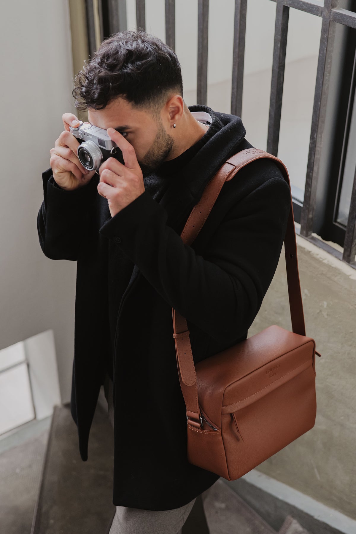 Man taking a photo with a camera, wearing a black coat and carrying a brown leather bag.