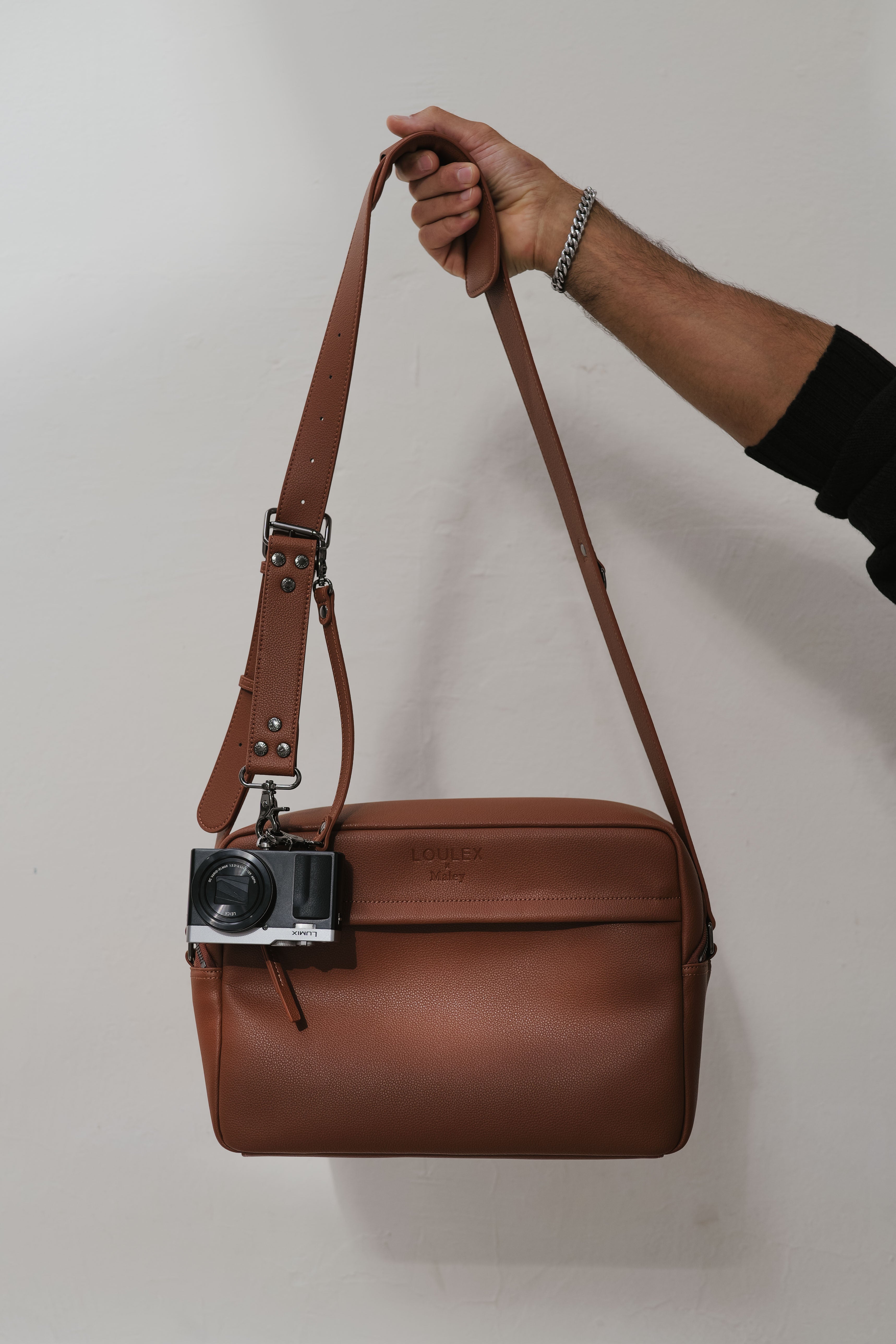 Brown leather camera bag with a vintage camera attached to it, held by a hand against a plain background.