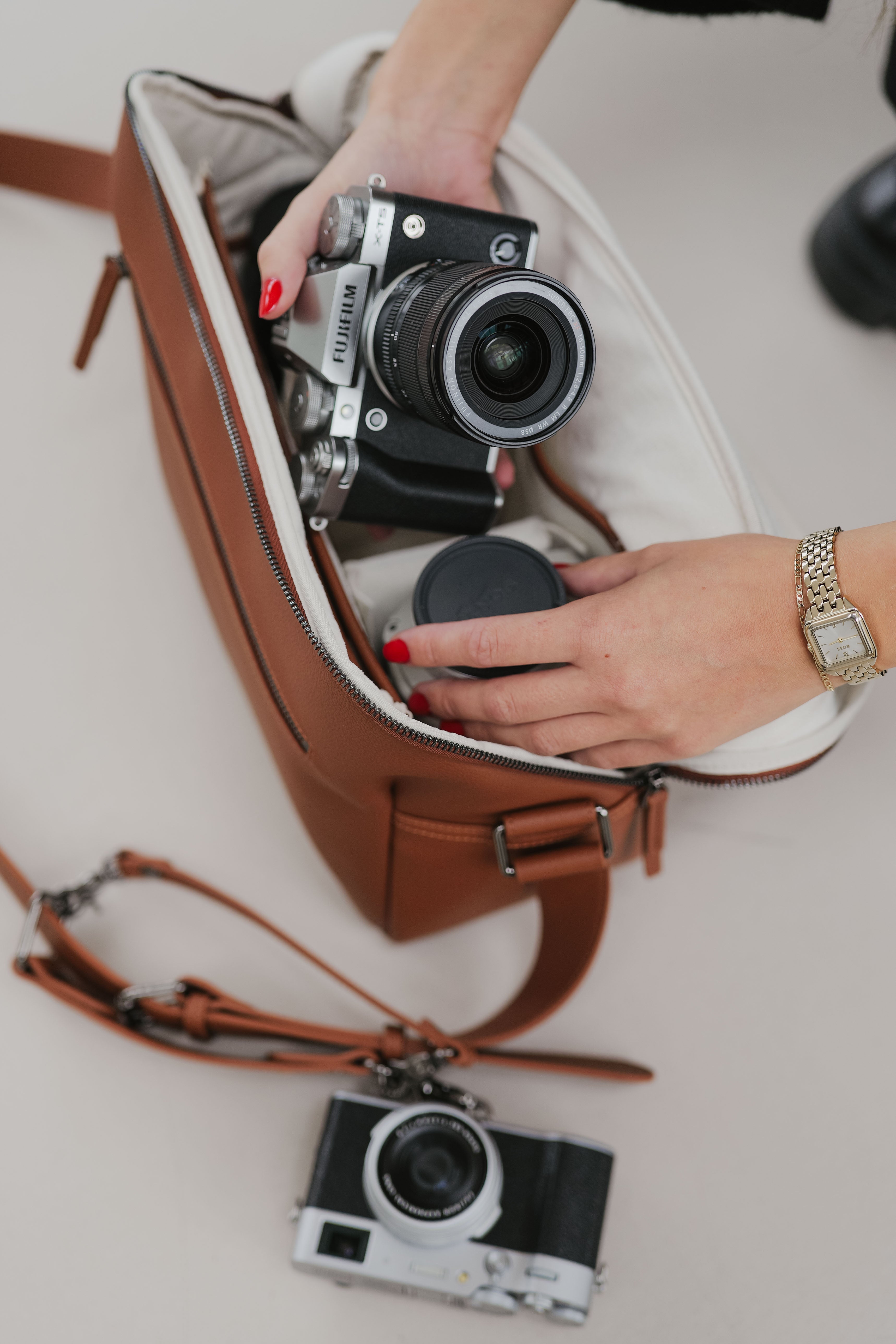 Person organizing a camera in a brown leather bag with another camera on a white surface.