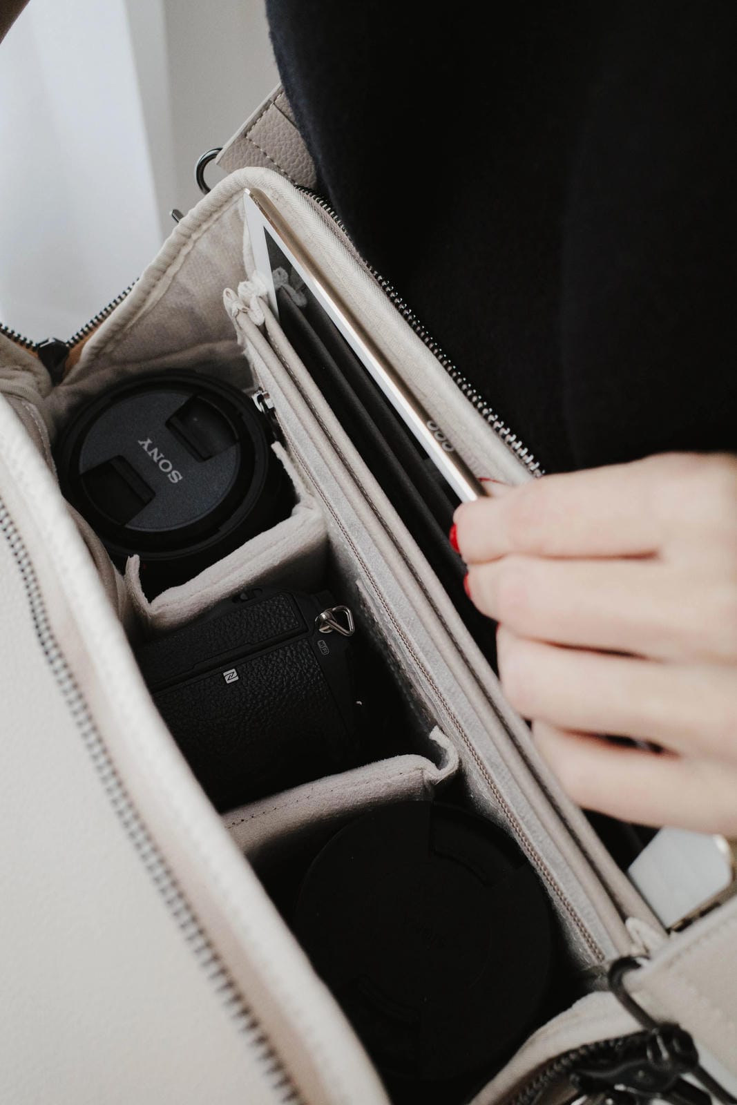 Hand holding a beige travel bag with compartments, one of which contains a black camera lens.